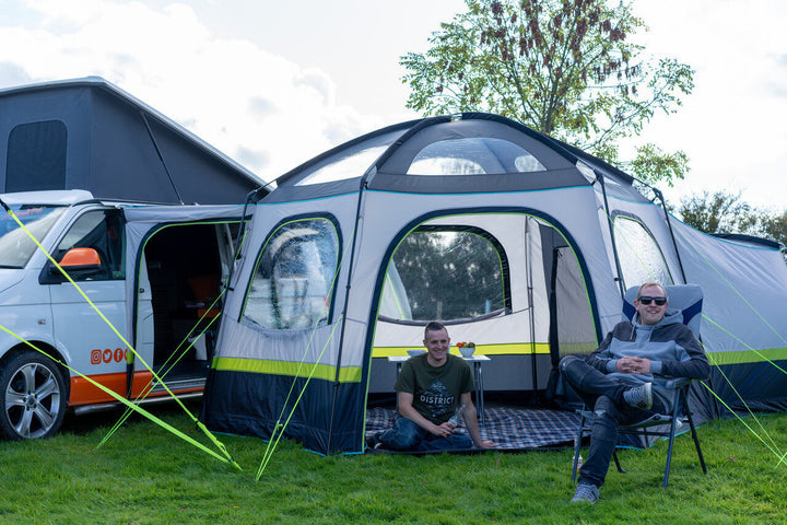 OLPRO Hive Campervan Awning set up beside a campervan with two men relaxing on chairs on a grassy field.