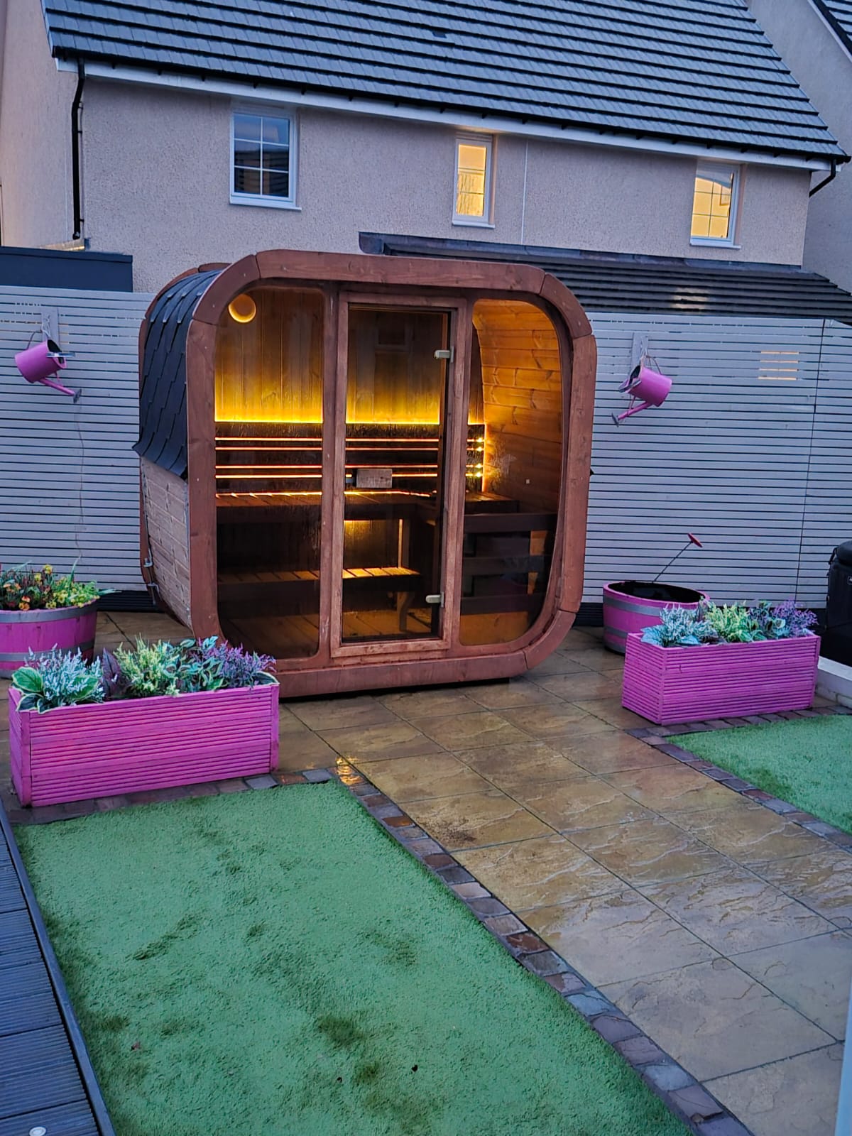 Wooden outdoor Icon 125 sauna with pink planters and green grass in a residential setting