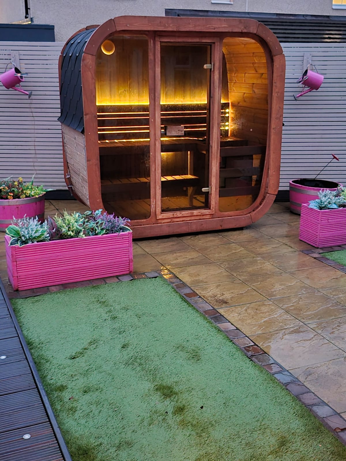 Wooden outdoor Icon 125 sauna with pink planters and a green mat on a patio.