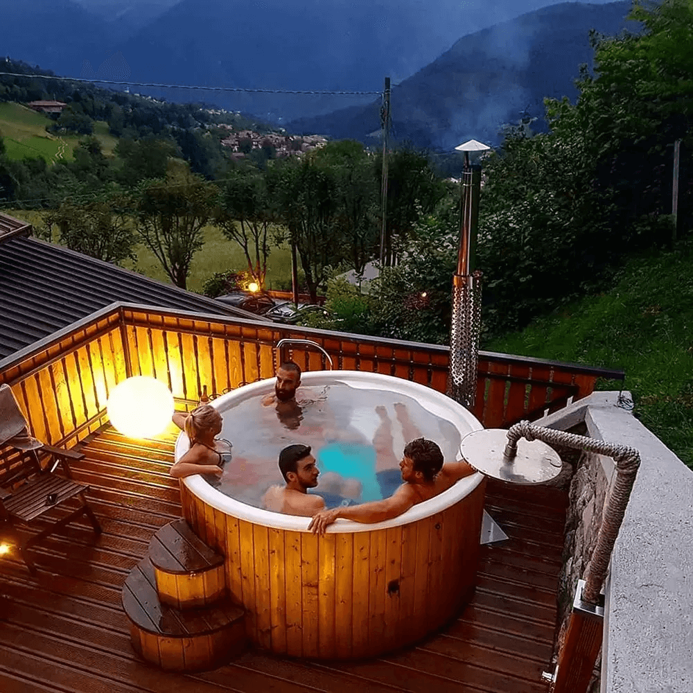 Group of people relaxing in an InMedias Delux 200 Hot Tub at dusk, on a mountain-view wooden terrace with ambient lighting.