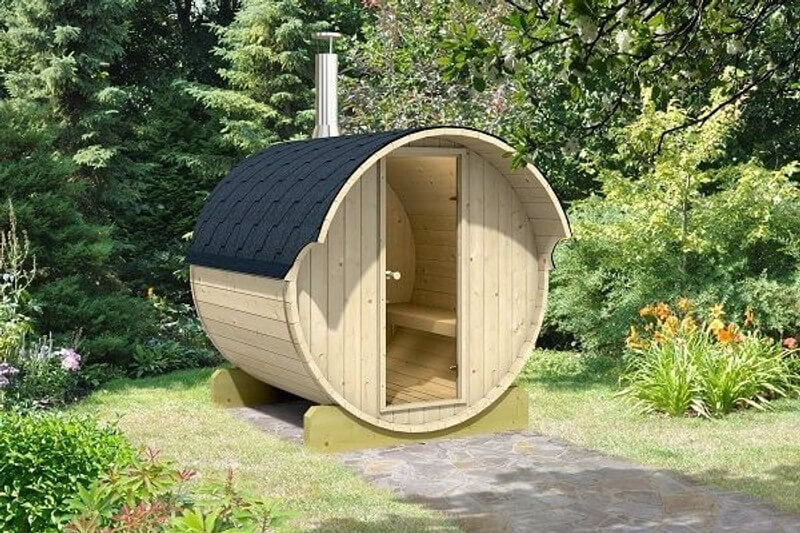 InMedias Barrel Sauna 220 placed on a garden stone path surrounded by green foliage and flowering plants, with a chimney extending from the roof.
