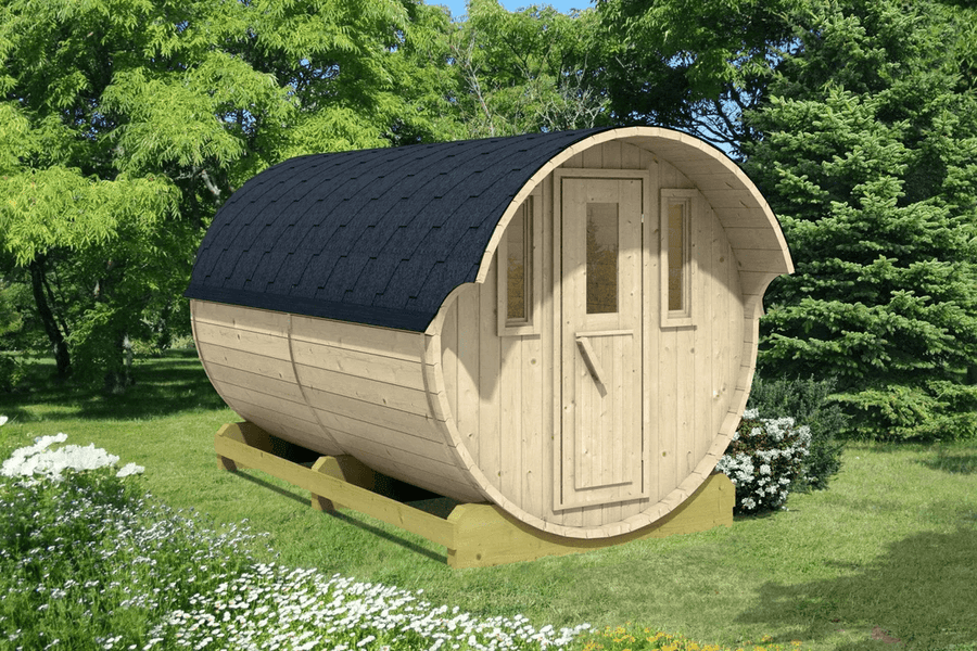 A wooden barrel cabin with black shingles, set in a lush green garden with white flowers and dense trees in the background, on a sunny day.