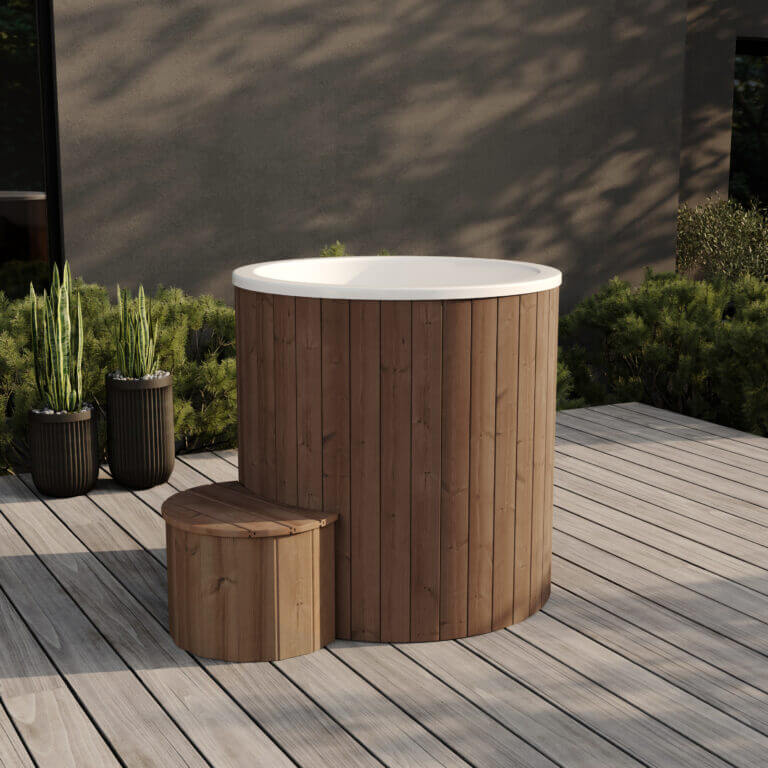 White interior cold tub with vertical brown wooden exterior and small semi-circular step, placed on a wooden patio with green bushes and tall planters.