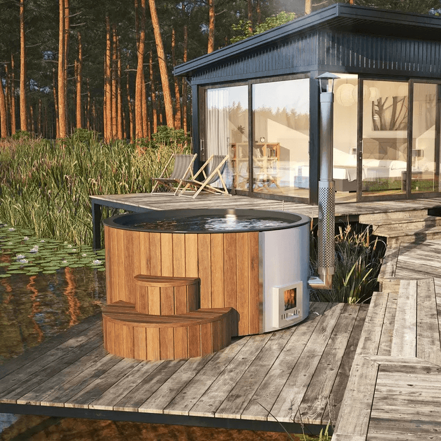 InMedias Delux 220 XL Hot Tub on a rustic wooden deck beside a modern cabin near a lake, surrounded by reeds and forest in daylight.