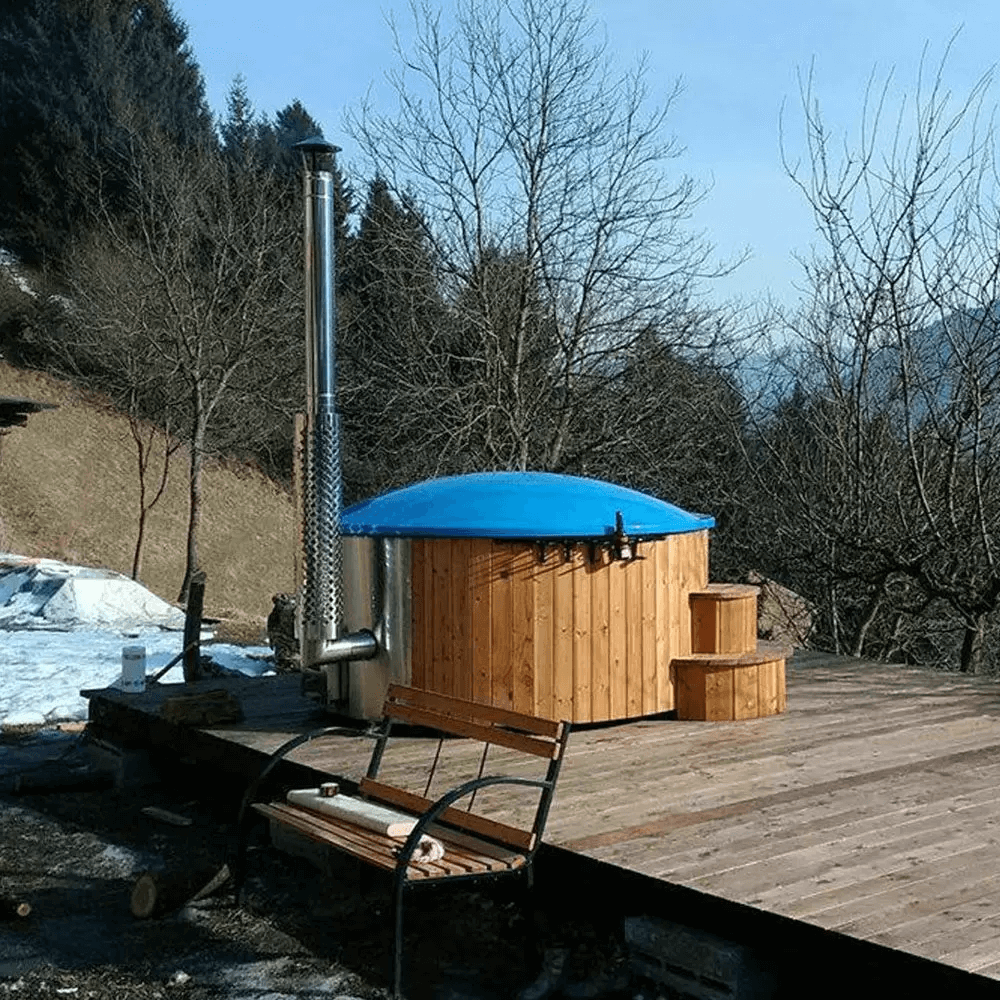 Wood-fired InMedias Delux 200 Hot Tub with blue lid and stainless steel chimney, placed on a wooden deck in a snowy mountain landscape.