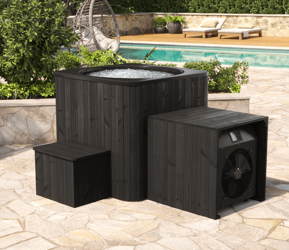 Black square cold tub with bubbling water, pump box, and wooden steps beside a swimming pool on a tiled patio.