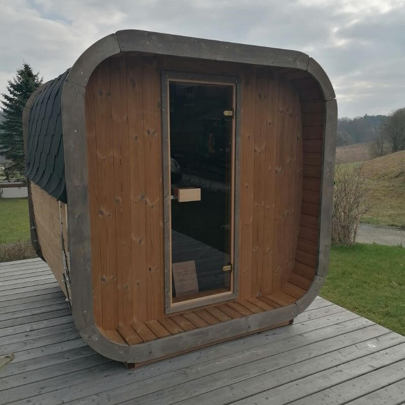 InMedias Sauna Icon 200S with dark wood front and black shingle roof, placed on a grey wooden platform under cloudy sky.