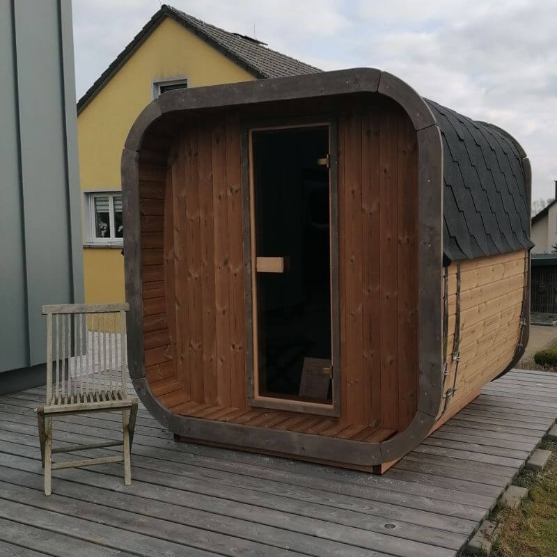 InMedias Sauna Icon 200S with rustic wood tones beside a yellow house, captured on a cloudy day.