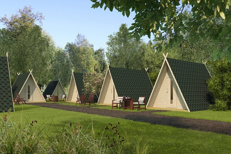 InMedias Wigwam Glamping Pod – exterior view of multiple A-frame cabins in a grassy outdoor setting with green shingle roofs and light wood doors, surrounded by trees and outdoor furniture, on a natural background.