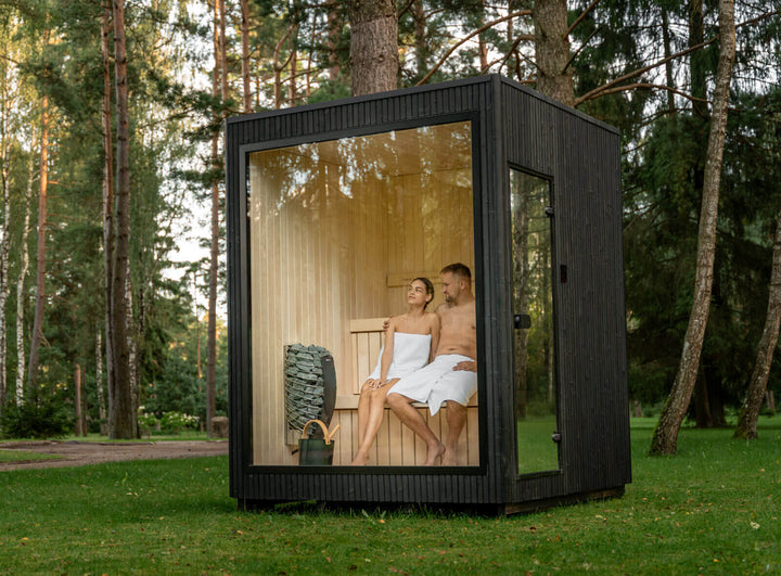 Jonas Classic Outdoor Sauna ebony finish with couple seated inside, located on green lawn in forest.