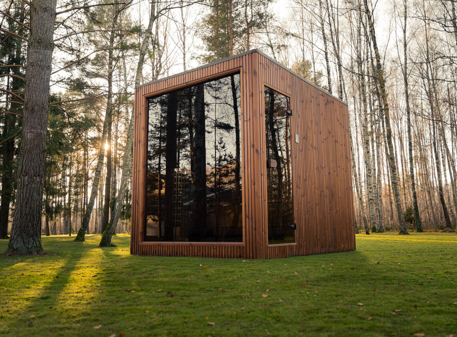 Jonas Grand Classic Outdoor Sauna exterior in teak finish with full-height tinted glass façade, installed on grass in a forest park at sunset.