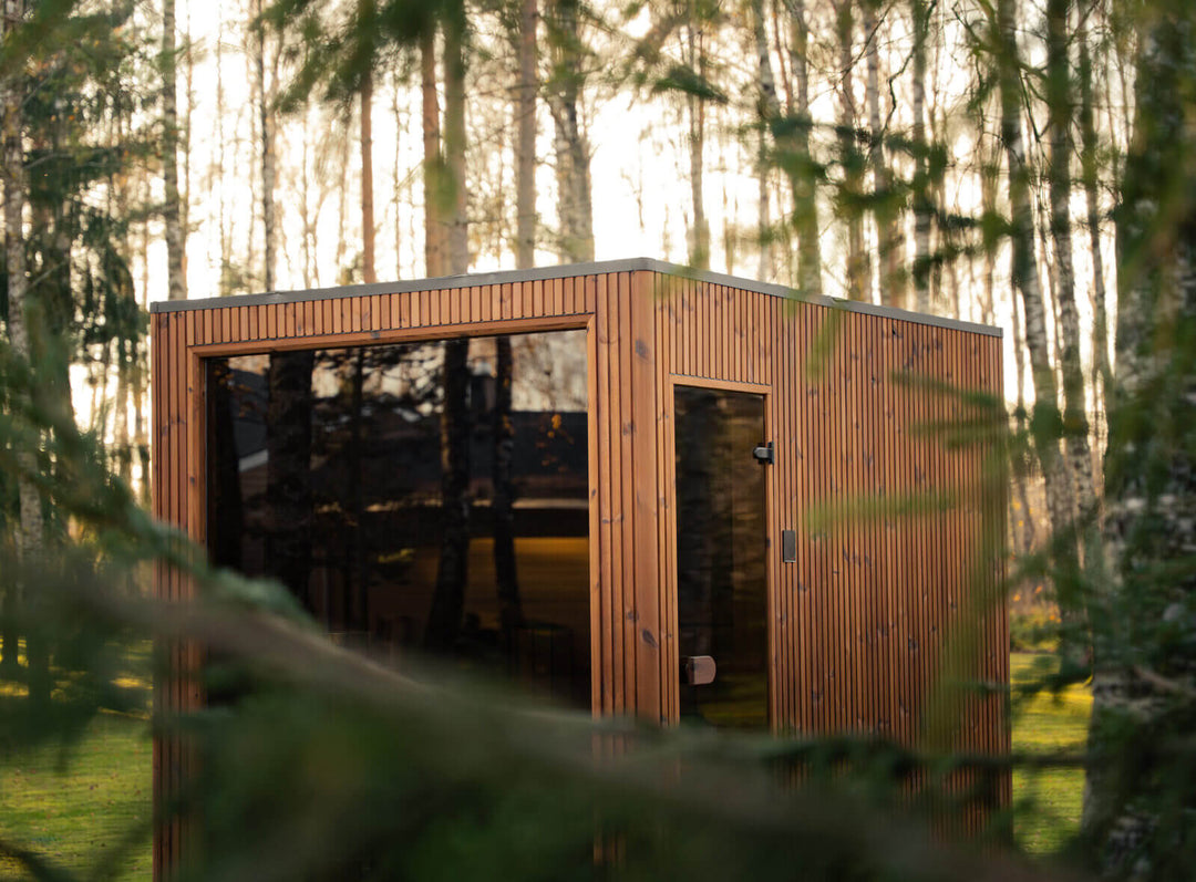 Jonas Grand Classic Outdoor Sauna in teak finish viewed through tree branches, featuring vertical timber cladding and dark glass panels in a woodland garden setting.