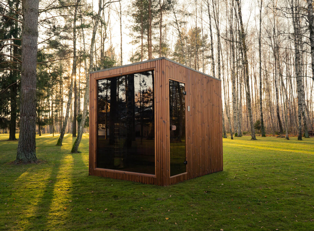 Jonas Grand Classic Outdoor Sauna corner view in teak finish with full-height tinted glass front, installed on grass surrounded by tall trees during golden hour.