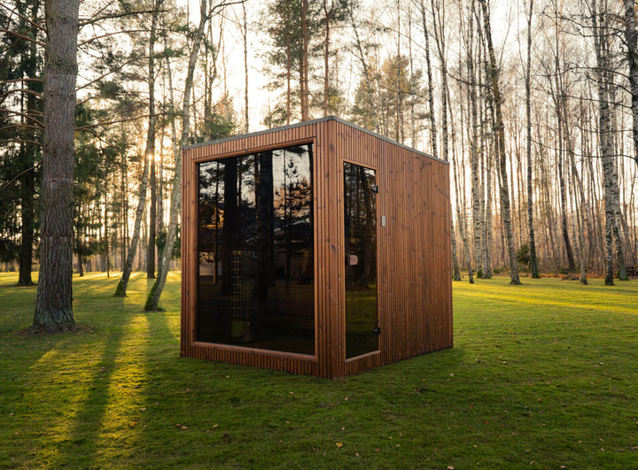Jonas Grand Classic Outdoor Sauna corner view in teak finish with full-height tinted glass front, installed on grass surrounded by tall trees during golden hour.