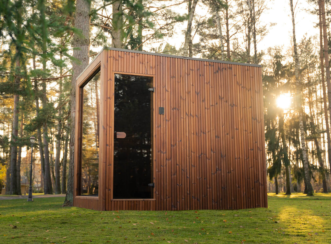 Jonas Grand Classic Outdoor Sauna in teak finish with vertical slatted wood exterior and tinted glass door, placed on a green lawn in a forest setting at sunset.