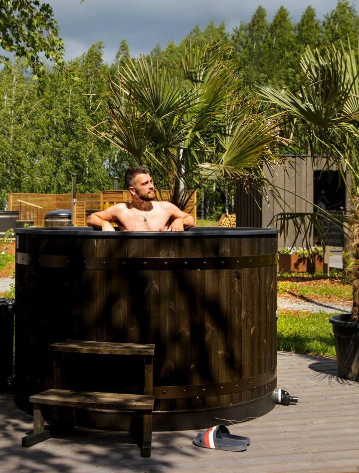 Man relaxing in a Harvia Legend black wooden hot tub outdoors surrounded by trees and garden decor.