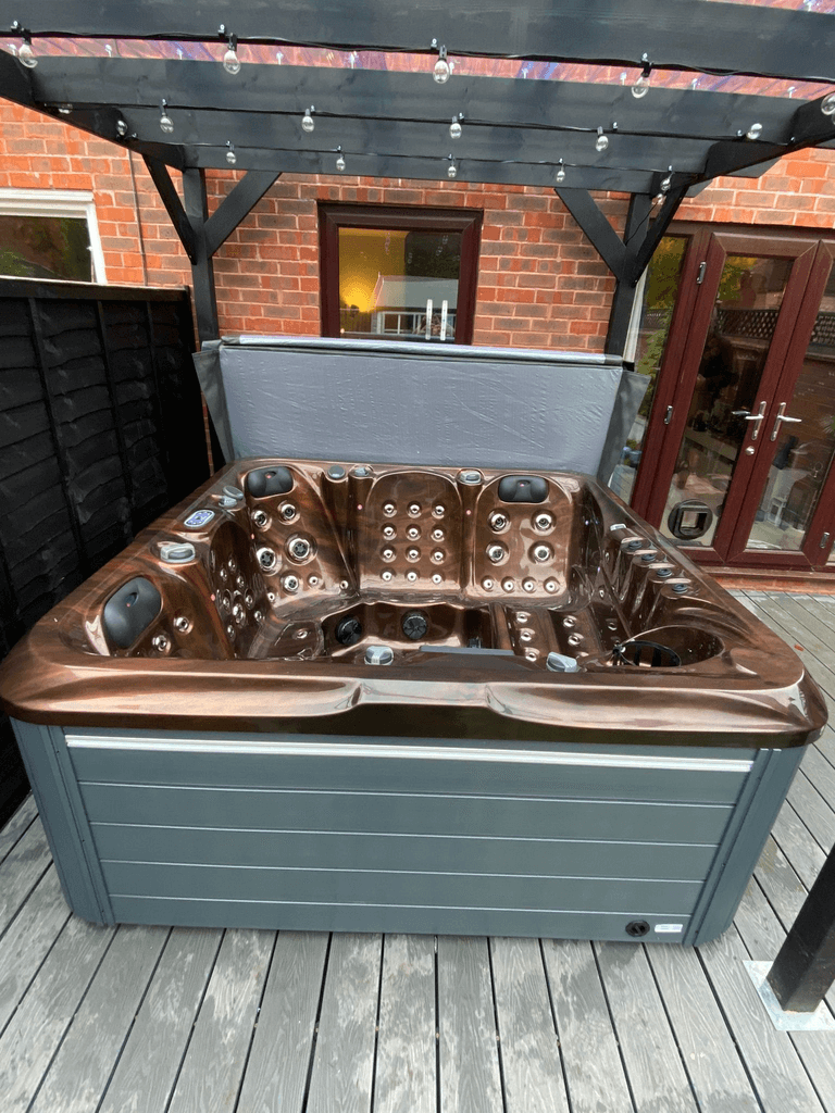 Luso Spas The 7000 Hot Tub in a brown marble finish installed outdoors under a pergola beside a brick wall and glass doors.