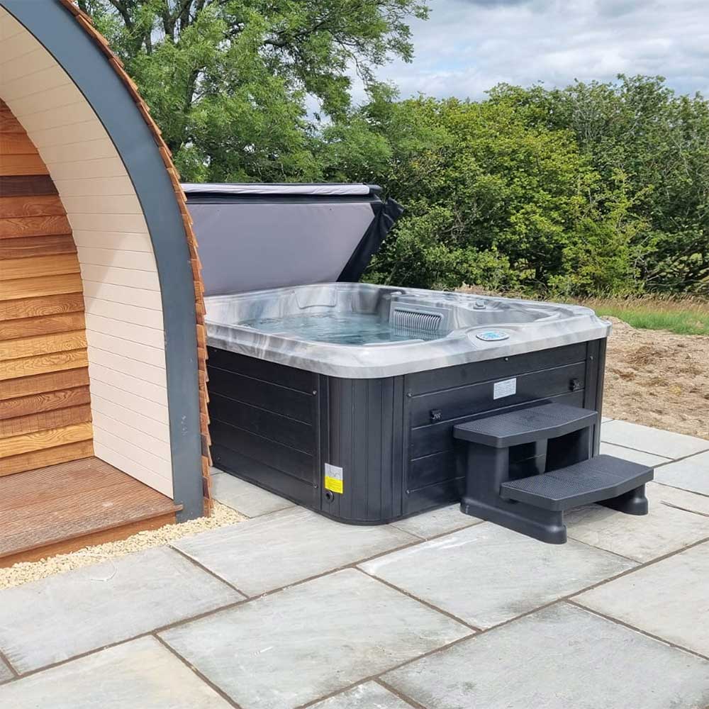 Outdoor installation of Luso Spas Sunbeach SB344S Hot Tub beside a wooden lodge, filled with water and fitted with black steps on a stone patio.