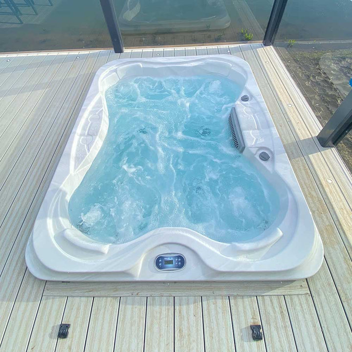 Top-down view of Luso Spas Sunbeach SB344S Hot Tub in white acrylic finish, filled with bubbling water on a wooden deck beside glass panels.