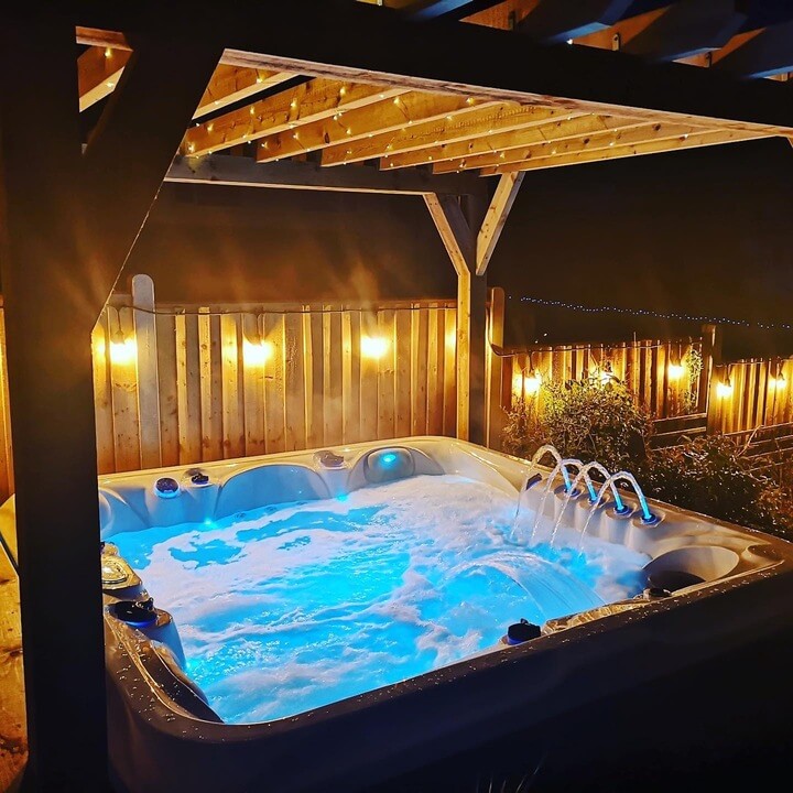 Luso Spas The 7000 Hot Tub illuminated with blue LED lighting under a wooden pergola at night, surrounded by warm ambient lights.