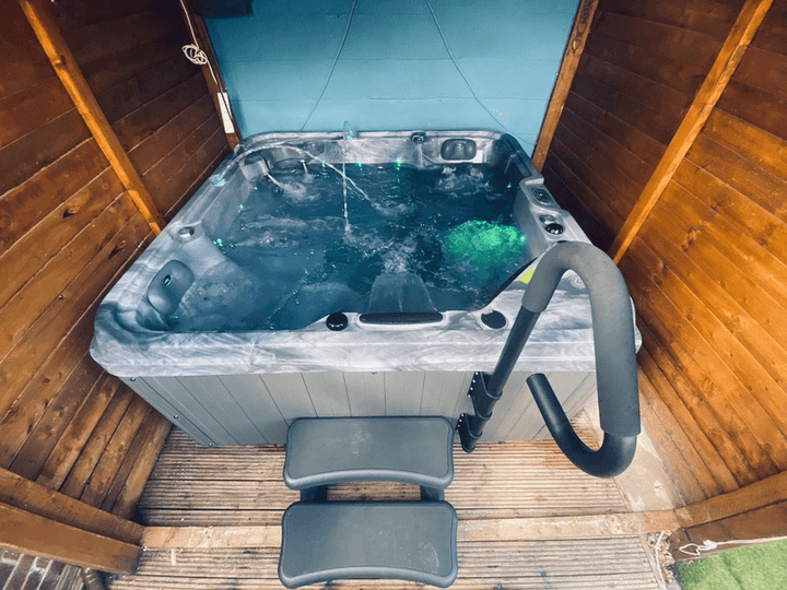 Luso Spas The Hawaii 2 Hot Tub bubbling with green LED lights in a wooden enclosure on a deck with metal safety rail and steps.