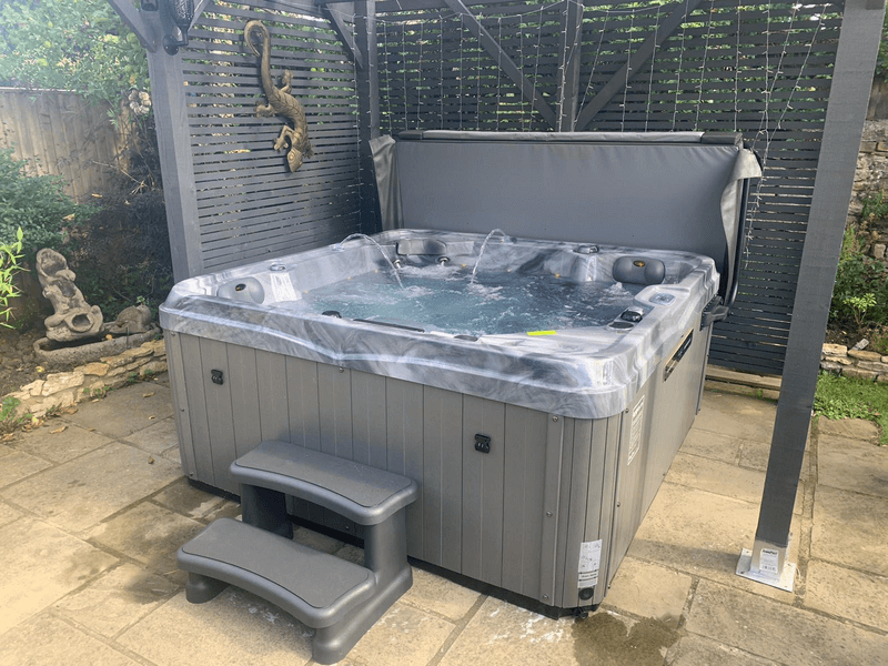 Luso Spas The Hawaii 2 Hot Tub bubbling in a paved garden area under a grey pergola with decorative elements in the background.