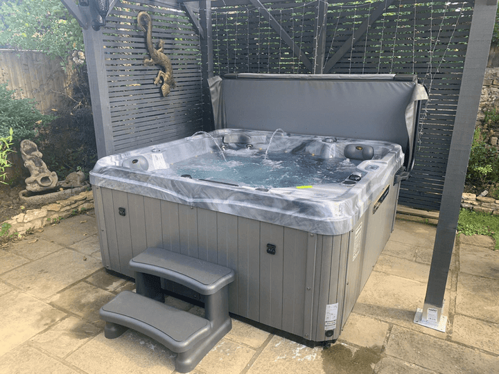 Luso Spas The Hawaii 2 Hot Tub bubbling in a paved garden area under a grey pergola with decorative elements in the background.