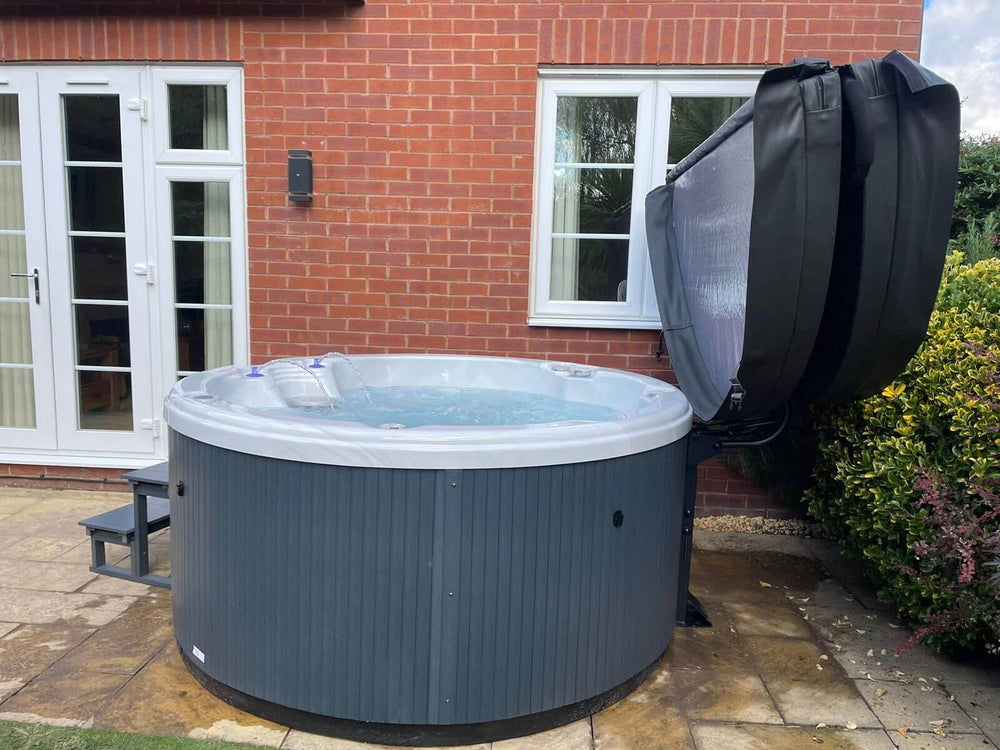 Outdoor view of Luso Spas The Zodiac Plug & Play Hot Tub placed beside a red brick house with open cover and bubbling water.
