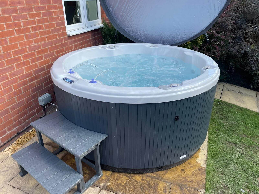 Angled view of Luso Spas The Zodiac Plug & Play Hot Tub beside a house wall with open insulated cover and grey entry steps.