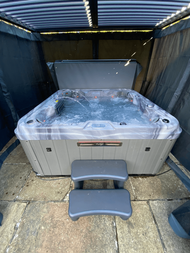 Front view of Luso Spas The Venice Hot Tub with cover partially open, bubbling water, and steps positioned in front under a pergola.