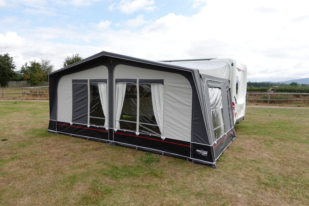 Camptech Mayfair DL Seasonal Caravan Awning with clear windows and white curtains, attached to a white caravan on a grassy field with trees and hills in the background.