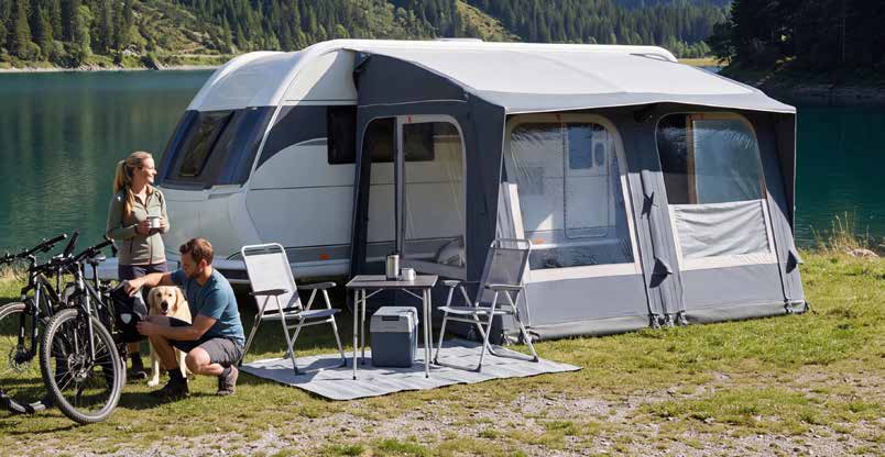 A large gray and white Starcamp Navigator Air 260 caravan awning with an attached outdoor dining set is pitched beside a lake, with two people and a dog sitting nearby on a grassy camping site.