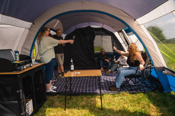 People relaxing inside the OLPRO Cocoon V2 awning with furniture and camping gear, set up on grass.