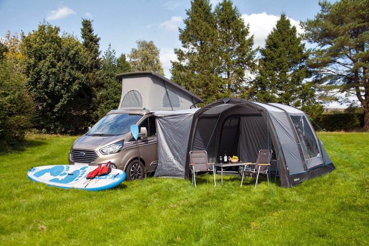 Wide campsite view of the Outdoor Revolution Cayman Air T3 Lite driveaway awning connected to a campervan with a paddleboard and outdoor seating.