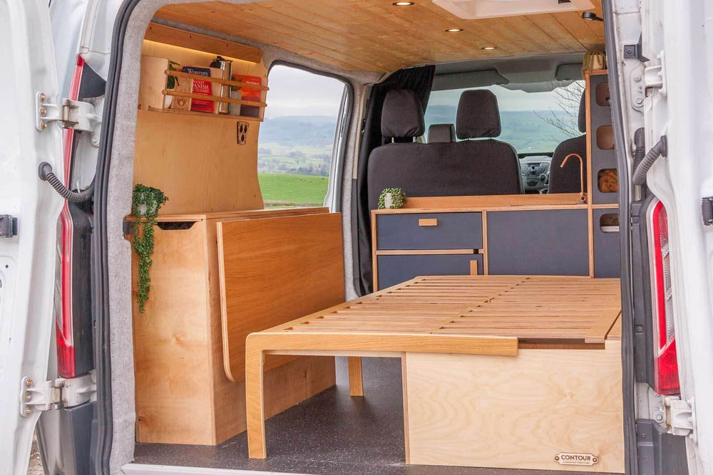 The Campervan Rig Sideboard is shown installed along the side of the van, featuring a clean oak finish with contrasting cupboard fronts and a slatted pull-out bed system extended across the rear space.