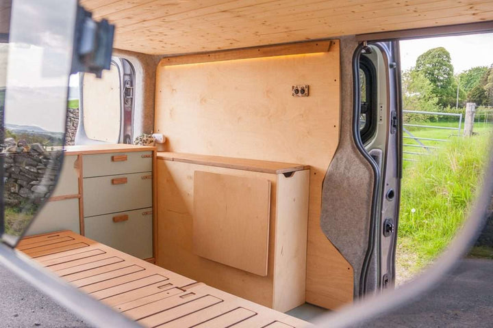 Campervan Rigette Sideboard with fold-down table folded away against the wooden side unit, viewed through the van window with grassy countryside outside.