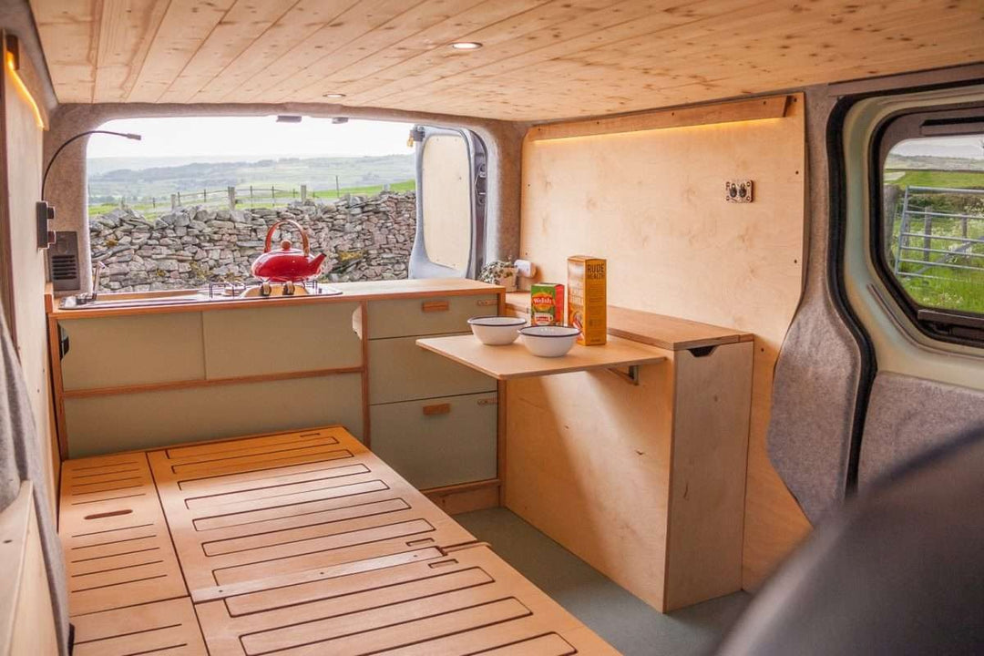 Campervan Rigette Sideboard setup with fold-down table extended and wooden cabinetry along the side wall, inside a campervan parked in a rural countryside setting.