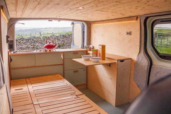 Campervan Rigette Sideboard setup with fold-down table extended and wooden cabinetry along the side wall, inside a campervan parked in a rural countryside setting.