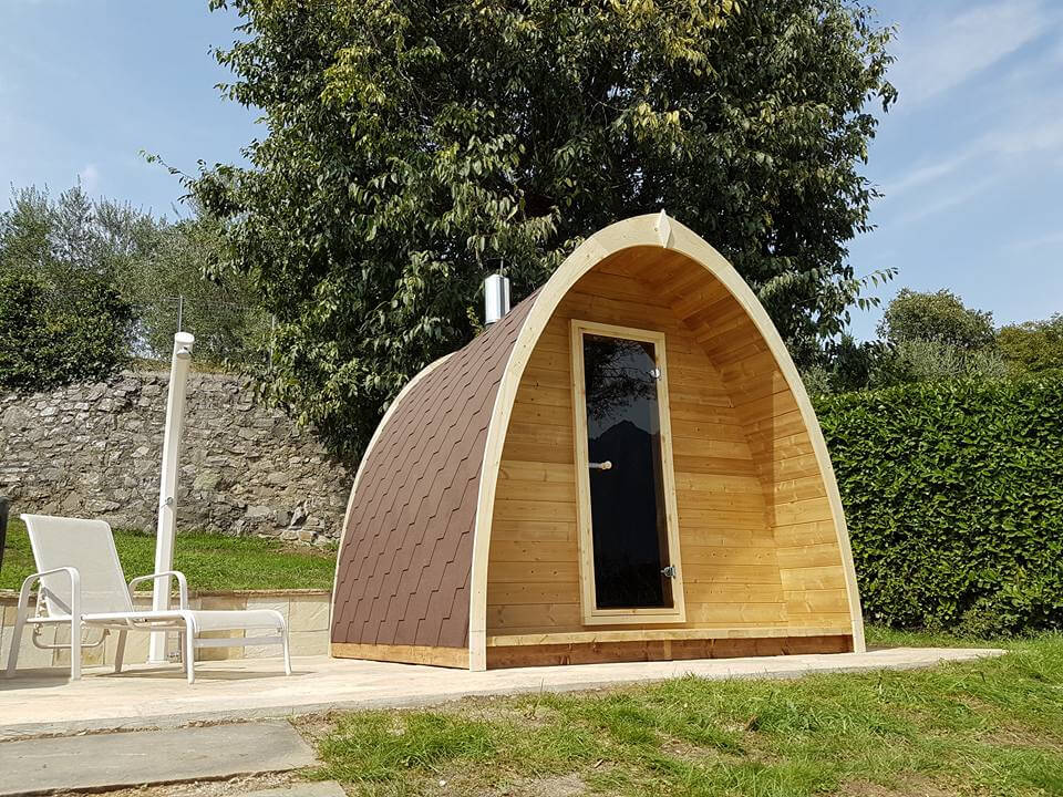 InMedias Sauna Pod 300 viewed from the front right angle wooden decking and vertical handle glass door on a sunny day with greenery in background.