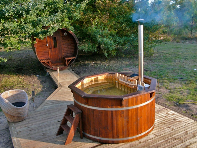 Dark wood Viking Industrier Spruce hot tub with steam rising, placed on a wooden deck surrounded by trees and a barrel sauna in the background.