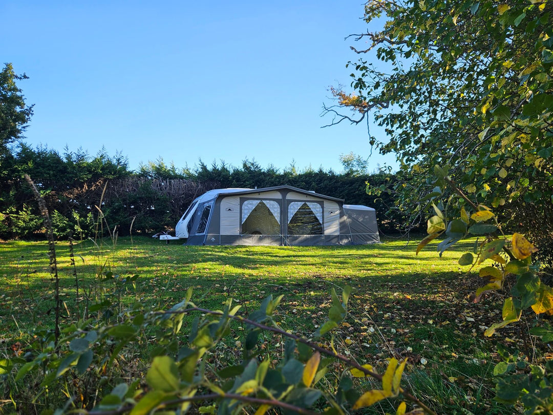 Wide view of the Sunncamp Casa Full Touring Poled Awning attached to a caravan on a grassy field surrounded by trees and bushes under a clear blue sky.