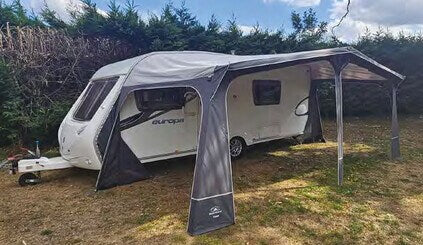 Sunncamp Casa Full Touring Poled Awning set up beside a white touring caravan on a grassy campsite with hedges and cloudy sky in the background.