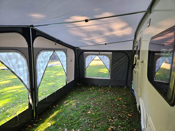 Interior view of the Sunncamp Casa Full Touring Poled Awning attached to a caravan with window panels, grass floor, and trees visible outside in the background.