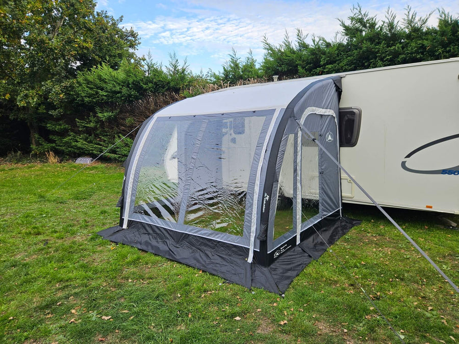Front view of the Sunncamp Ultima Versara Air 260 Deluxe Porch Awning with large panoramic windows attached to a caravan on a grassy campsite with trees and hedges.