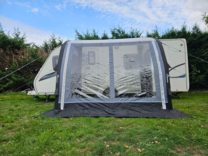 Side view of the Sunncamp Ultima Versara Air 260 Deluxe Porch Awning connected to a white caravan on grass with hedge trees and cloudy sky.