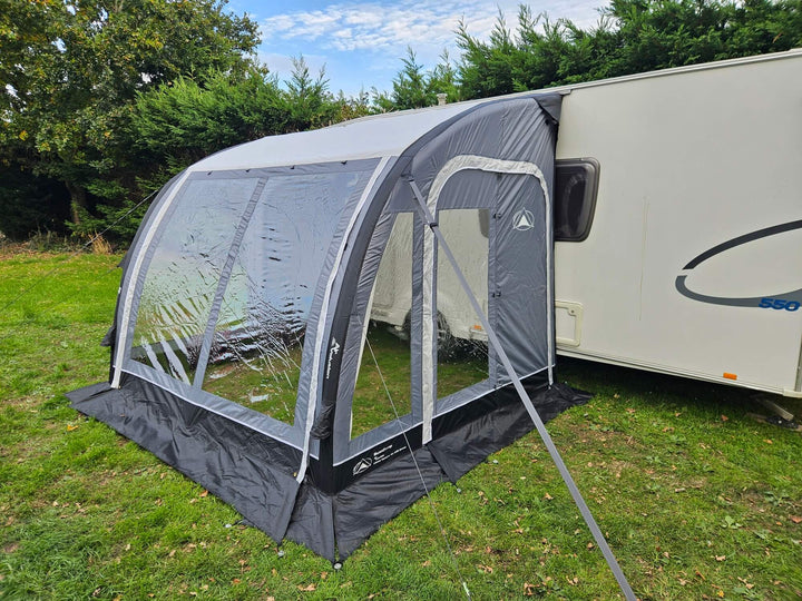 Front view of the Sunncamp Ultima Versara Air 260 Deluxe Porch Awning with window covers closed attached to a caravan on a grassy campsite with hedge trees and cloudy sky.