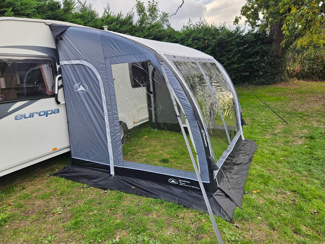Front angled view of the Sunncamp Ultima Versara Air 260 Deluxe Porch Awning attached to a caravan with clear window panels on a grassy campsite with trees and hedges.