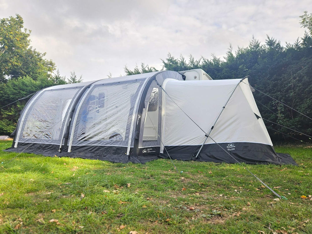 Side view of the Sunncamp Ultima Versara Air 390 Deluxe Porch Awning with rear annex extension attached to a caravan on a grassy campsite with hedges in the background.