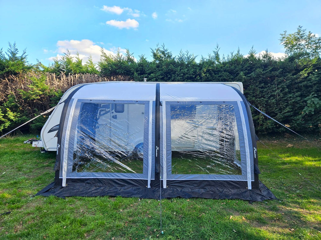 Front view of the Sunncamp Ultima Versara Air 390 Deluxe Porch Awning with large clear windows set up beside a caravan on grass with hedge trees and blue sky in the background.