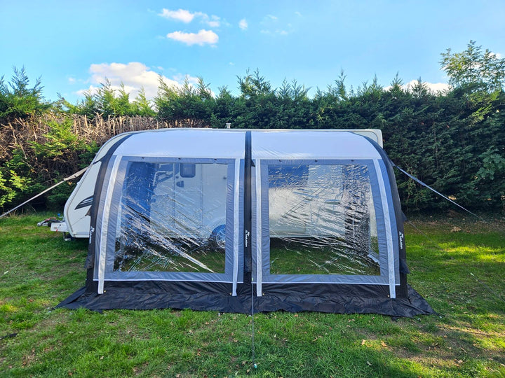 Front view of the Sunncamp Ultima Versara Air 390 Deluxe Porch Awning with large clear windows set up beside a caravan on grass with hedge trees and blue sky in the background.